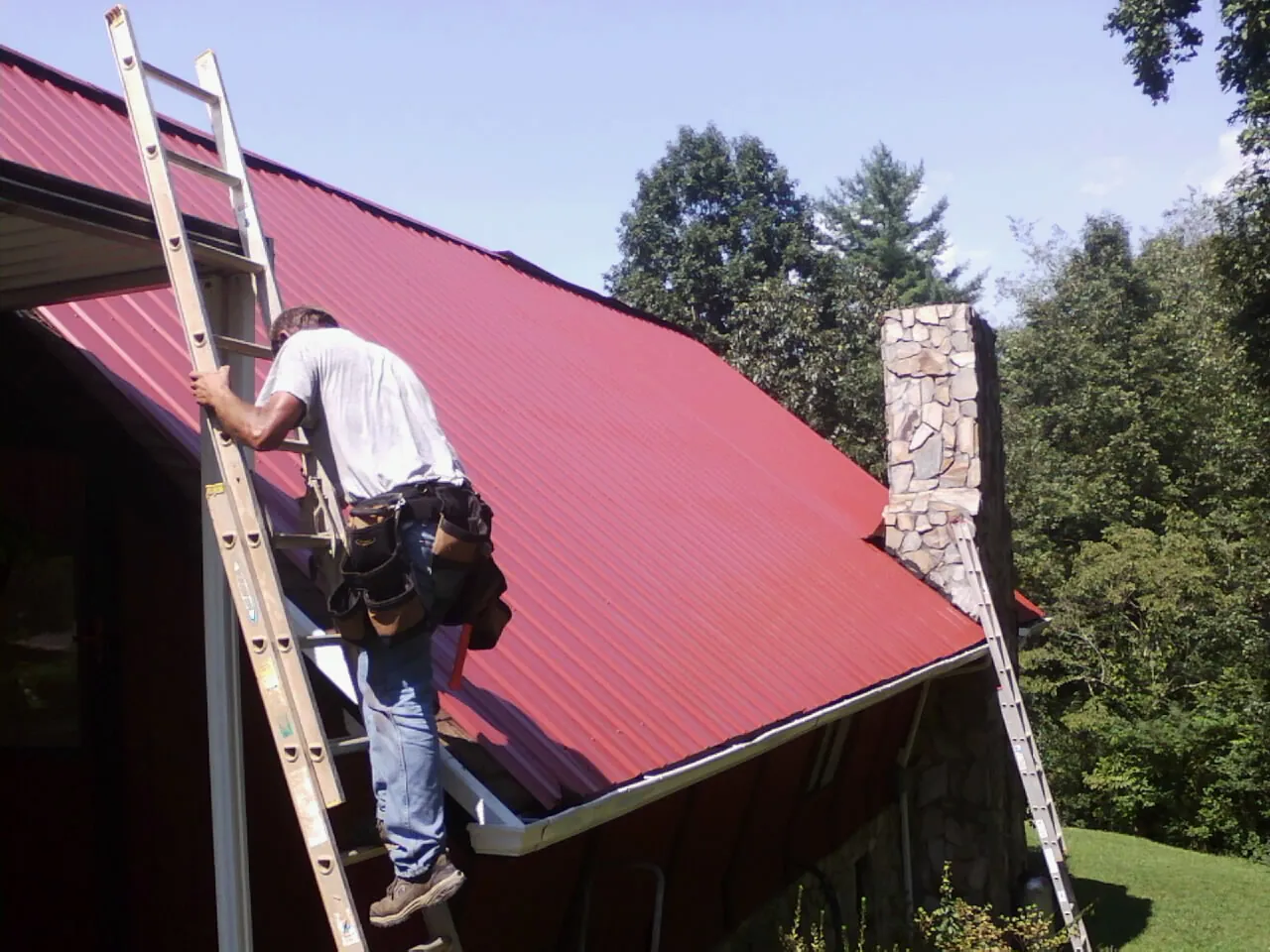 Roofer on a ladder beside a red metal roof and stone chimney — Soffit & Fascia Repair by Roofing Brothers in Union Hall